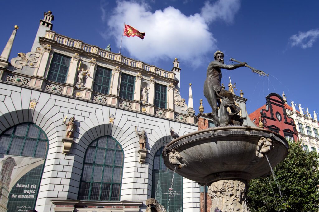 A grand building with arched windows, stone carvings, and a central statue atop a fountain. A red and yellow flag waves above against a blue sky.