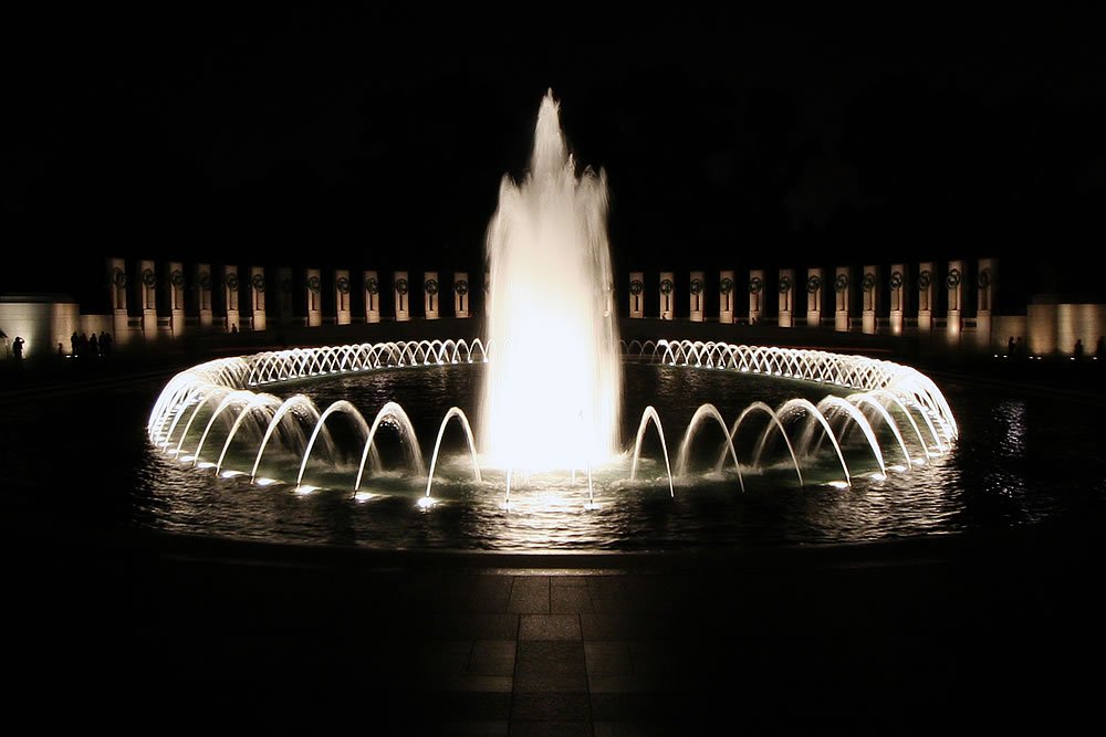 A bright, glowing fountain illuminated at night, with water arching gracefully into the circular basin. The surrounding area features softly lit columns in the background.