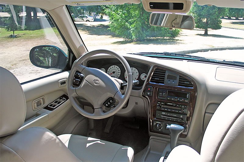 This image shows the front interior of a 1990s Lexus car with a beige dashboard, wood trim accents, and an automatic transmission. The driver’s seat comes into view, and the side window reveals a natural outdoor scene.