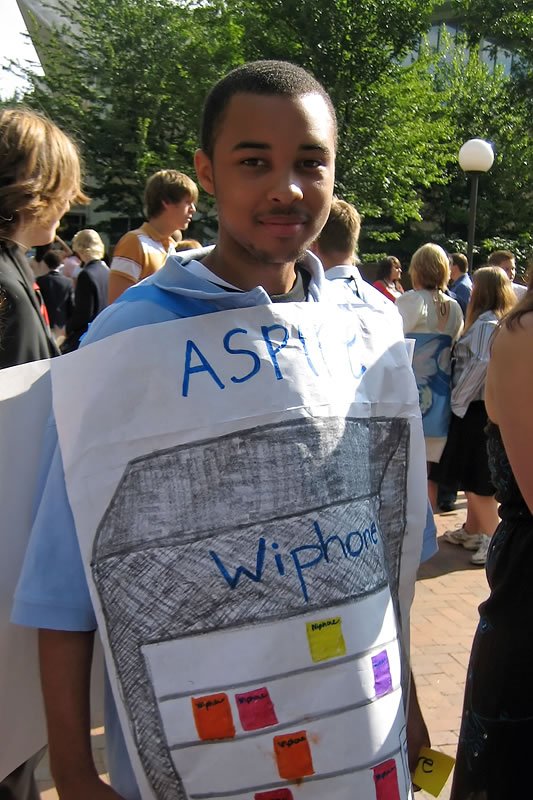 A young man wearing a homemade costume resembling an iPhone, complete with hand-drawn screen details and app icons, at an outdoor gathering.