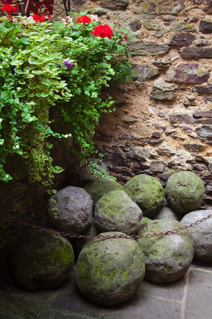 Four iron balls tied to a chain are used as an entryway security feature. They are next to old stone walls with red flowers growing nearby.