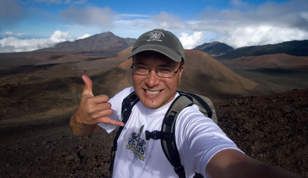 John taking a selfie with a thumbs-up gesture against a stunning volcanic landscape backdrop. The mountains feature varied textures and colors, under a partly cloudy sky.