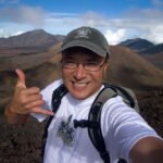 Big Beach & Little Beach John taking a selfie with a thumbs-up gesture against a stunning volcanic landscape backdrop. The mountains feature varied textures and colors, under a partly cloudy sky.