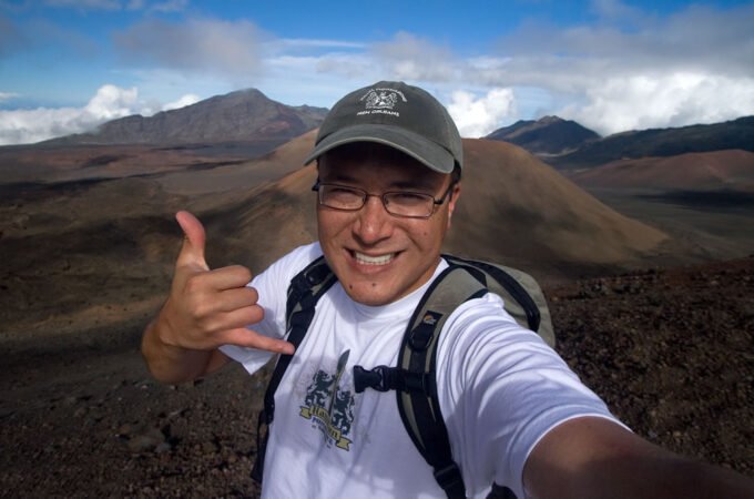 John taking a selfie with a thumbs-up gesture against a stunning volcanic landscape backdrop. The mountains feature varied textures and colors, under a partly cloudy sky.