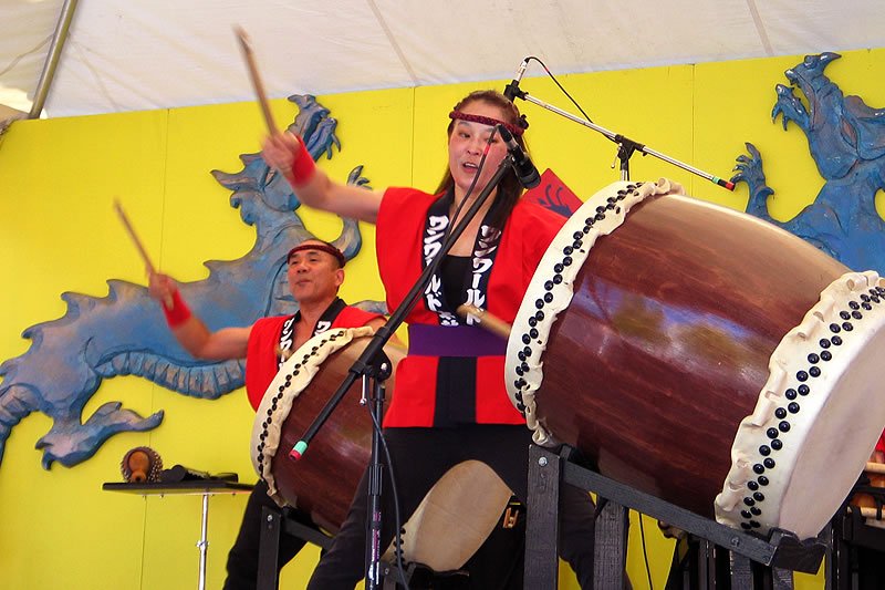Two performers energetically playing traditional Japanese taiko drums in front of a brightly colored backdrop.