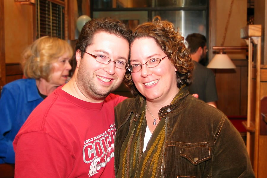 A smiling couple stands close together at an indoor gathering, surrounded by friends and cozy decor.