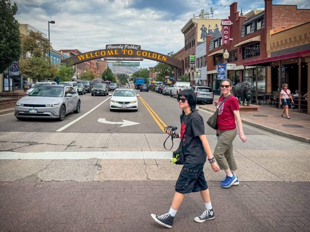 A couple walks across a city street beneath a large 'Welcome to Golden' archway. Cars drive by on the road lined with historic buildings.