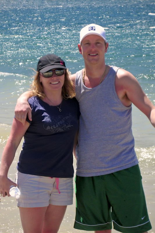 A smiling couple stands together near the water, both dressed in casual summer attire. The woman wears a navy blue tank top and light shorts, while the man is in a gray tank top and green shorts.