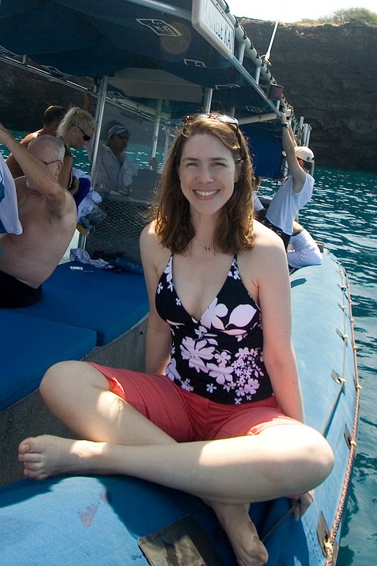 A happy woman in a floral swimsuit sits cross-legged on the edge of a blue boat, enjoying water views.