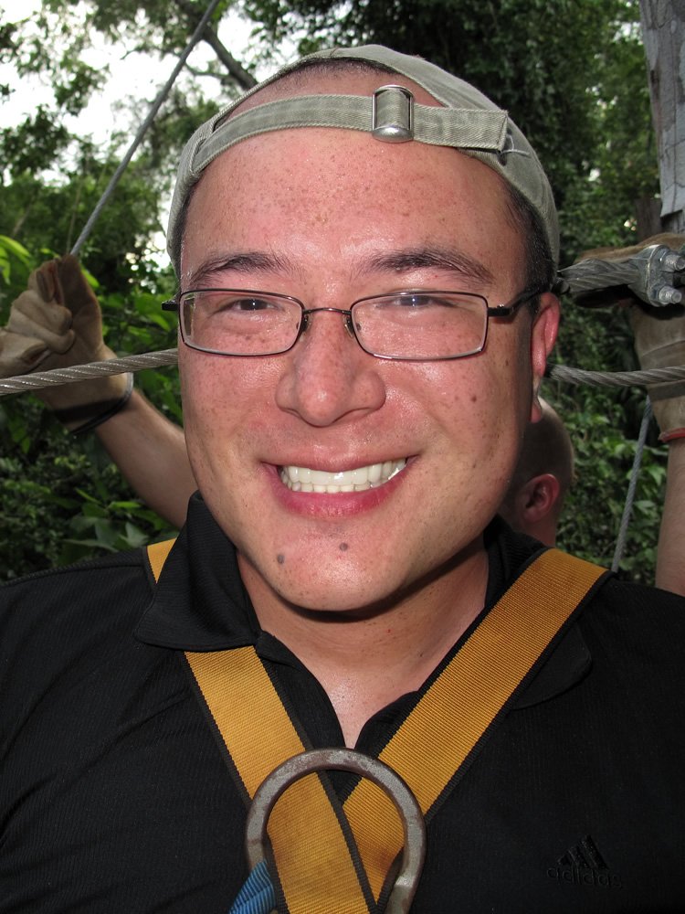 John with a headlamp and harness smiles during a zip-line excursion surrounded by lush greenery.
