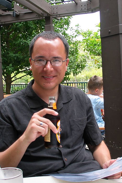 John holding a glass of beer, sitting at an outdoor table with greenery in the background.