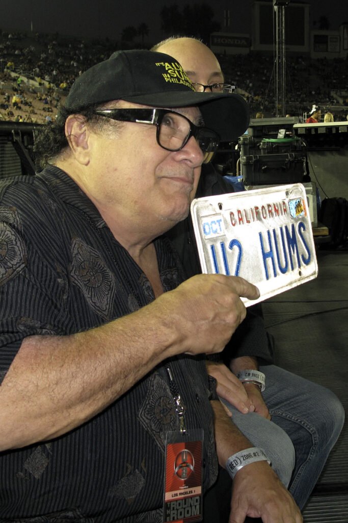 A person wearing a black hat sits at an outdoor event, proudly displaying a license plate that reads 'HUMS'. The setting appears casual and vibrant.