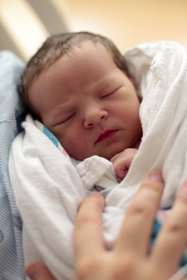 A serene newborn baby wrapped in a white blanket rests in a man's arms, his hand gently cradling the tiny infant. This intimate moment captures the new life and begins the love story for our family.