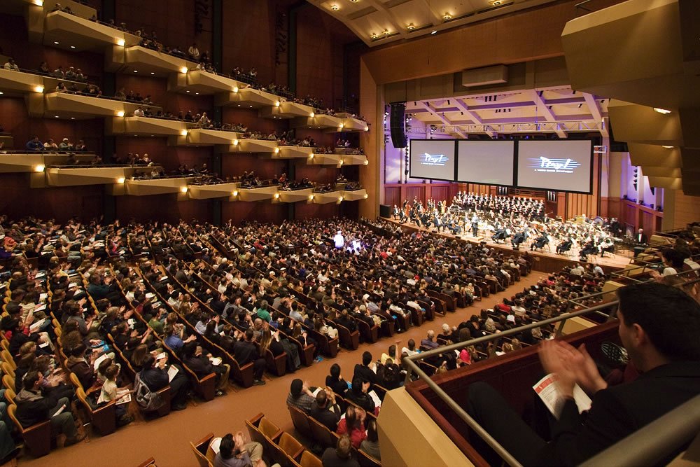 A large audience seated in a grand theater with multiple balconies, watching a presentation on stage screens.