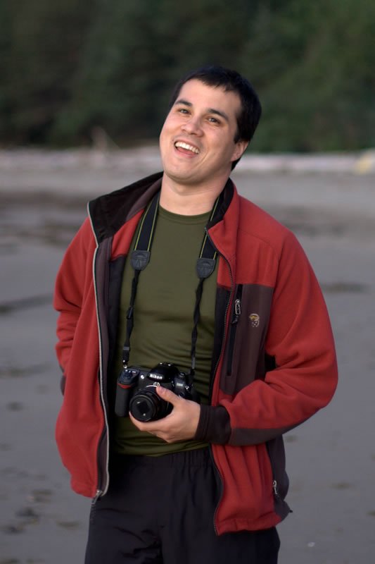 A man in a red jacket and green shirt laughs while holding a camera, standing on a beach near water and trees.