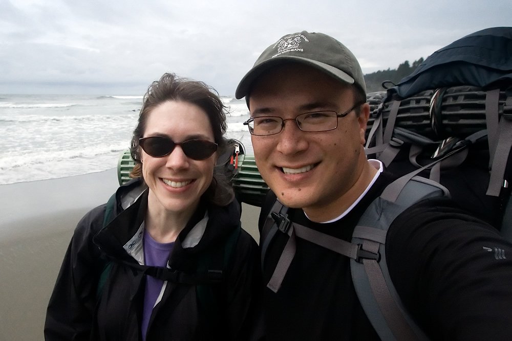 John and Kristie enjoy a sunny day at the beach. Both wear sunglasses and casual attire, ready for water activities.
