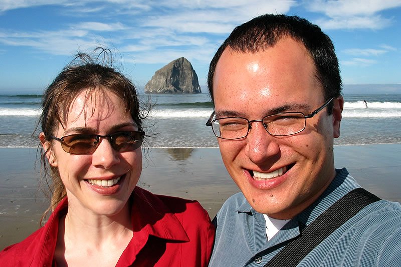 A couple smiling at the beach with Haystack Rock in the background.
