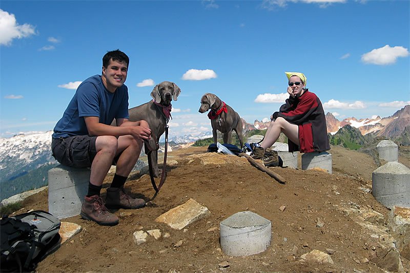 John and Kristie take a break on a mountain hike with their dogs. The scenic backdrop includes a clear blue sky and distant snowy peaks.