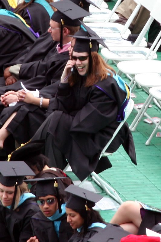 A candid photo of John and Kristie sitting among graduates at a graduation ceremony, dressed in black caps and gowns with teal accents. They are smiles and engaged in conversation amidst the bustling event.