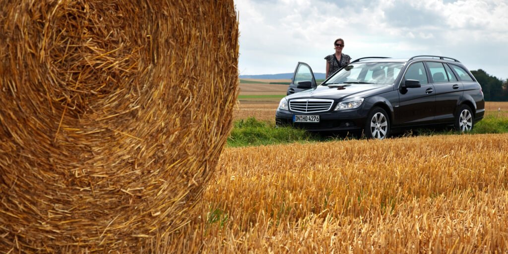 John and Kristie standing next to their car in a picturesque hayfield with a large hay bale nearby.