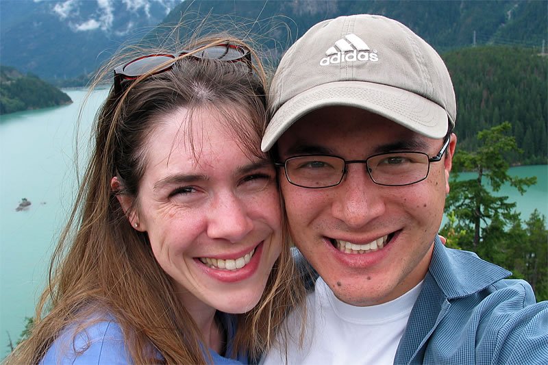 A couple, John and Kristie, smile joyfully while embracing against a stunning backdrop of a tranquil lake surrounded by lush mountains.
