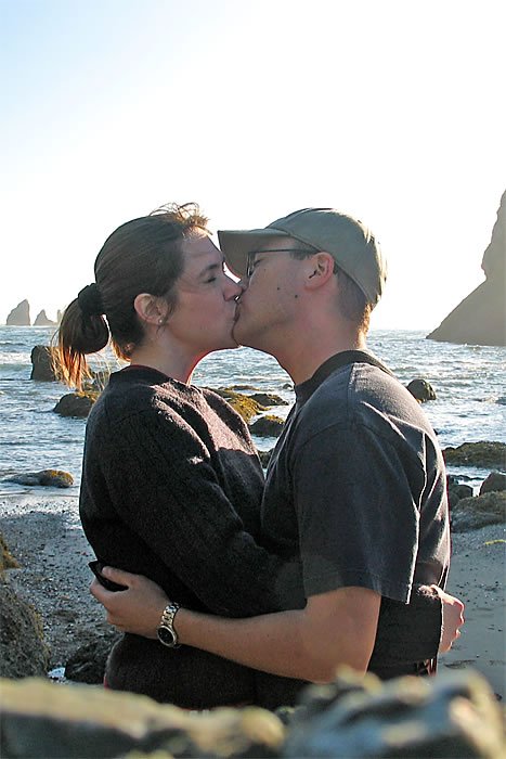 John and Kristie share an intimate kiss on a sunny day by the rocky Pacific Northwest shoreline.
