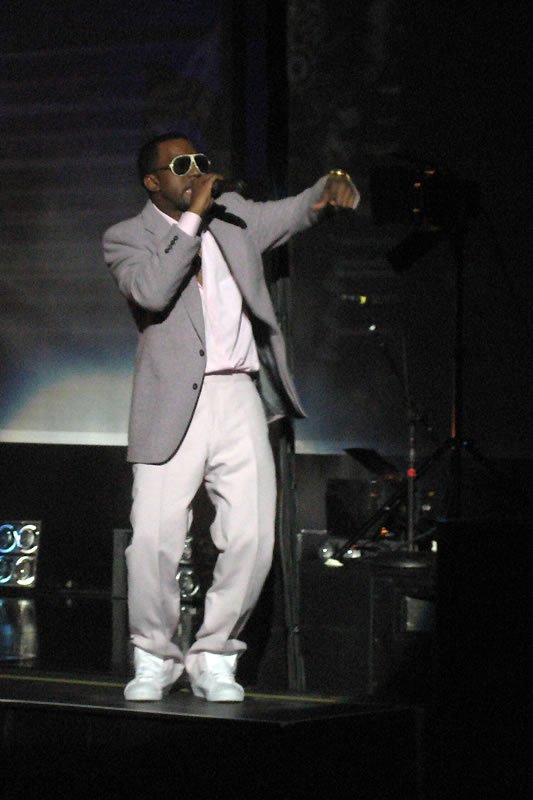 John stands on stage wearing sunglasses and a light grey suit, holding a microphone while performing at a concert.
