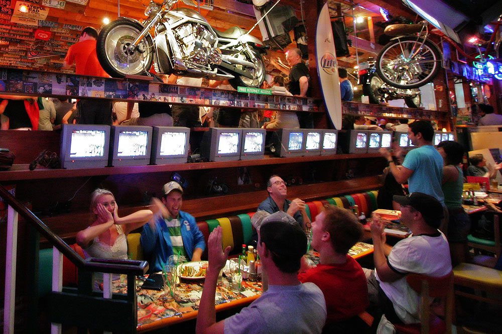 A group of people seated around a colorful table filled with dishes, chatting and laughing in a restaurant decorated with motorcycles and surfboards. The atmosphere is warm and vibrant, with wooden accents and ambient lighting enhancing the cozy setting.