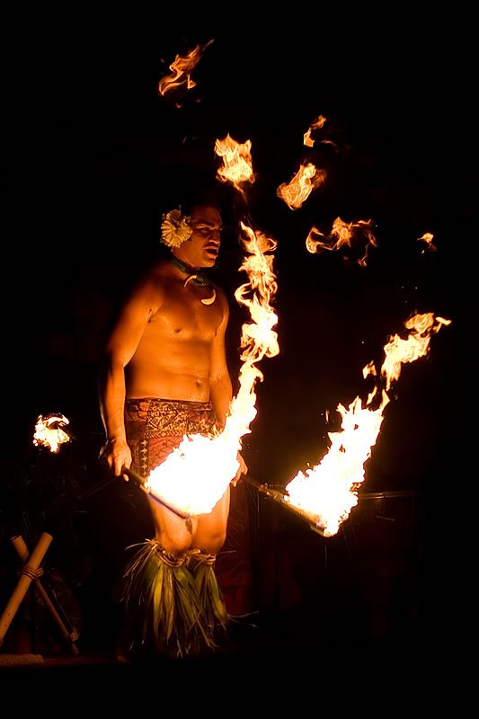 A shirtless dancer in traditional attire performs with flaming torches under the night sky.