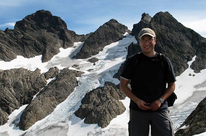 A person stands on a snowy mountain trail, surrounded by rugged peaks and clear blue skies. The image captures a close-up selfie with a scenic backdrop.