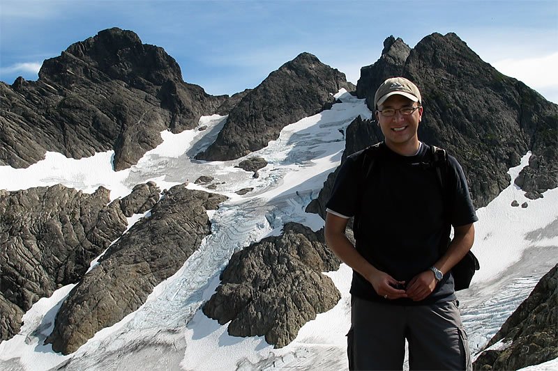 A person stands on a snowy mountain trail, surrounded by rugged peaks and clear blue skies. The image captures a close-up selfie with a scenic backdrop.