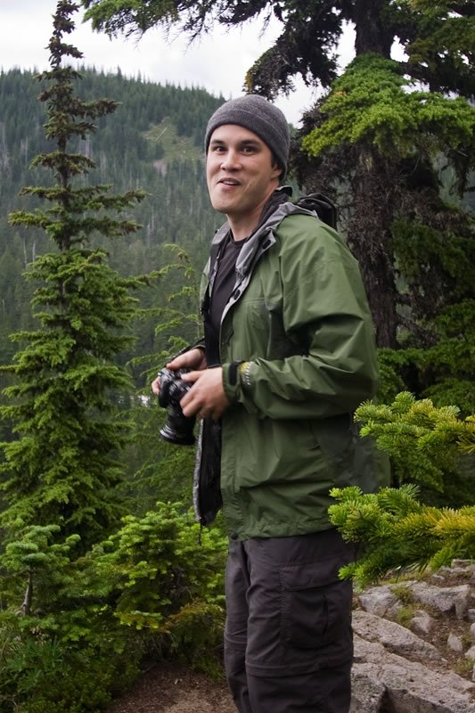 John stands in a forest holding a camera. The lush, green surroundings highlight our love for nature in the Pacific Northwest.