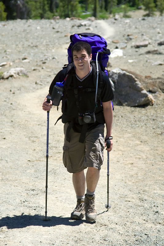A person with a purple backpack walking with hiking poles on a gravel path.