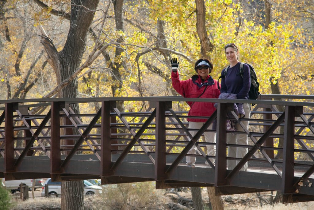 A couple, John and Kristie, stand on a bridge surrounded by vibrant yellow autumn leaves, with one of them raising a hand in wave.
