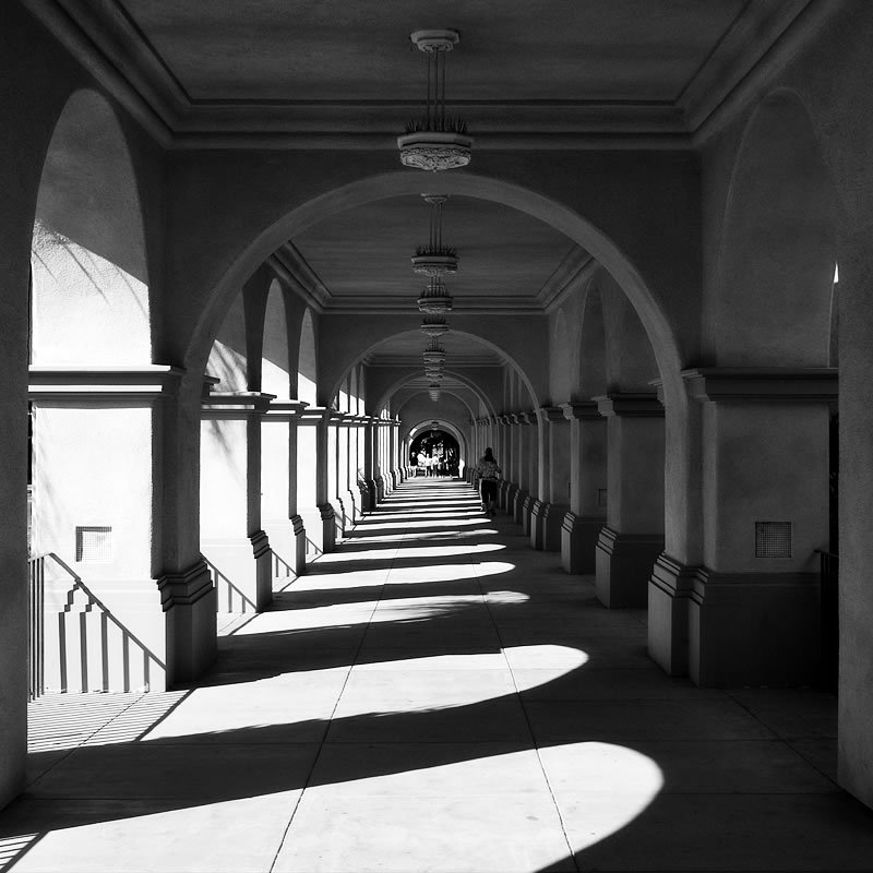 Black and white photo of John and Kristie walking hand-in-hand through arched corridors bathed in soft sunlight.