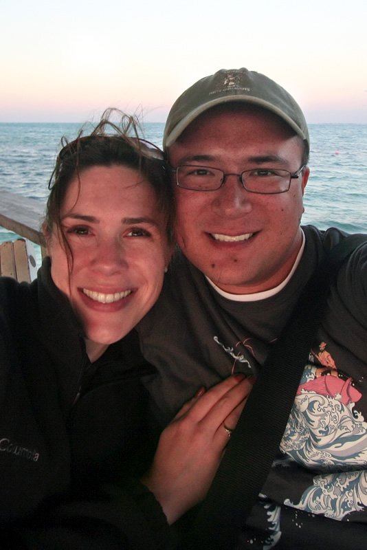 A close-up selfie of John and Kristie smiling on a boat. The background shows a calm sea under a pink sunset sky.