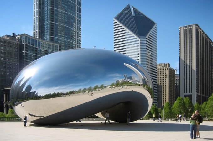 John and Kristie pose near the famous Chicago Cloud Gate sculpture, with skyscrapers in the background. The couple embraces by the shiny metallic structure on a sunny day.