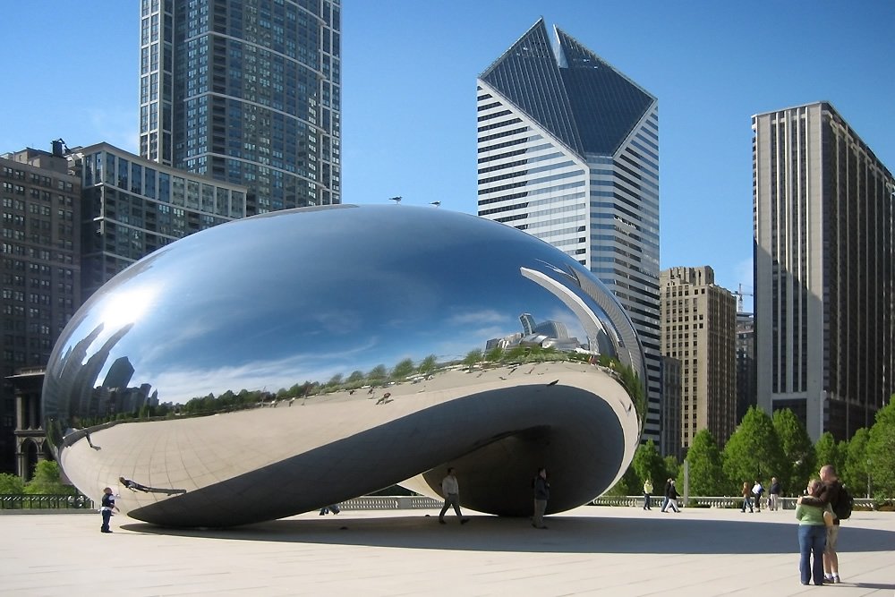 John and Kristie pose near the famous Chicago Cloud Gate sculpture, with skyscrapers in the background. The couple embraces by the shiny metallic structure on a sunny day.