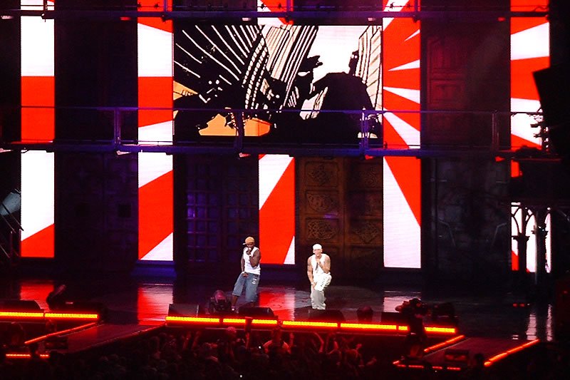Two performers on stage, illuminated by vibrant red and white lighting. The stage design features striking geometric patterns.