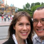 A smiling couple poses in front of Cinderella's Castle at a theme park. The iconic pink and blue castle stands majestically in the background.