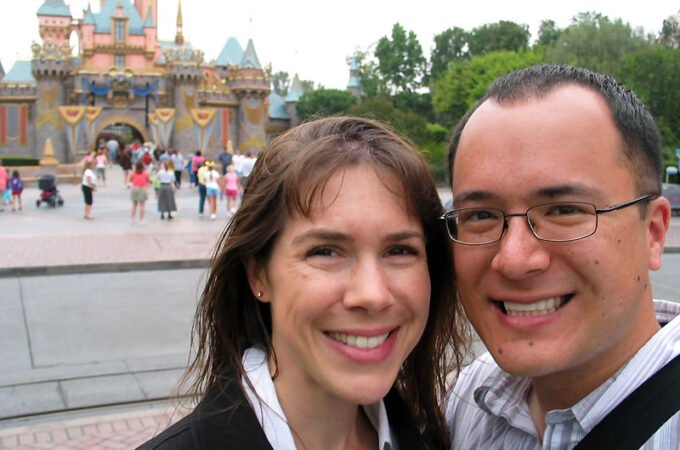 A smiling couple poses in front of Cinderella's Castle at a theme park. The iconic pink and blue castle stands majestically in the background.