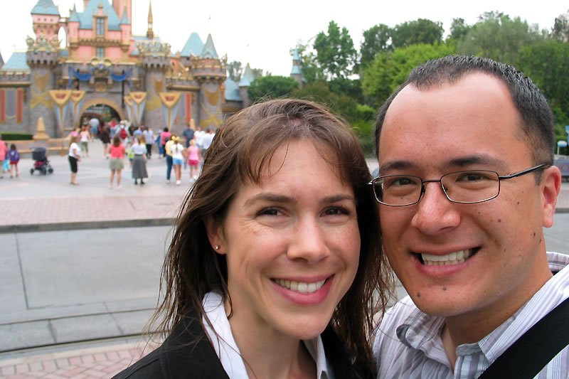 A smiling couple poses in front of Cinderella's Castle at a theme park. The iconic pink and blue castle stands majestically in the background.