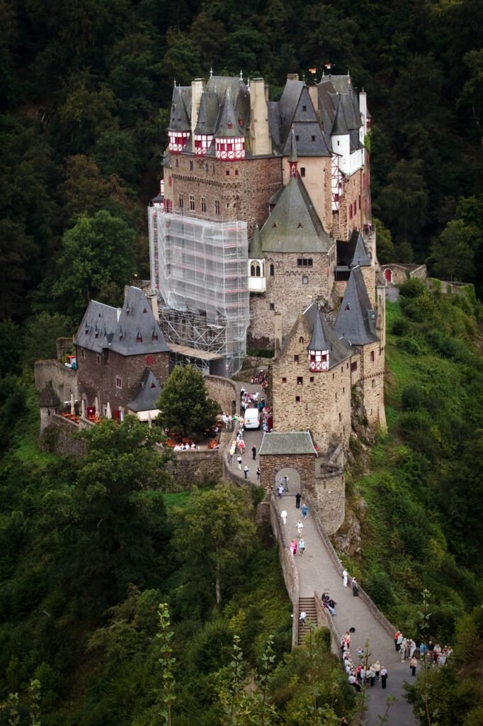 A majestic castle with steep slate roofs and red-tiled balconies rises dramatically from a lush green hillside, partially shrouded in mist. Tourists stroll along the winding road below as scaffolding clings to one tower, hinting at ongoing restoration.