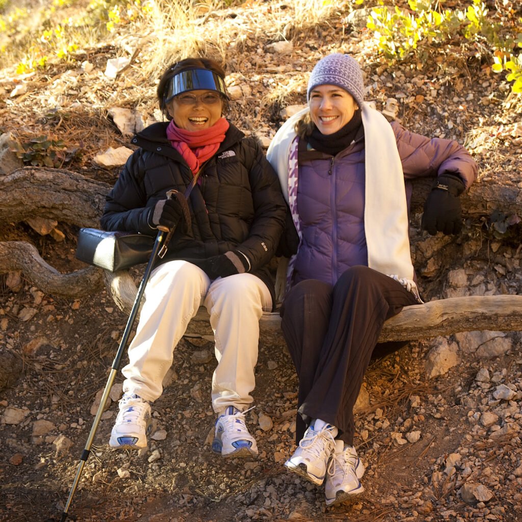 John and Kristie are sitting on a fallen log in a forest, dressed in winter hiking gear. They appear relaxed and happy, enjoying their outdoor adventure.