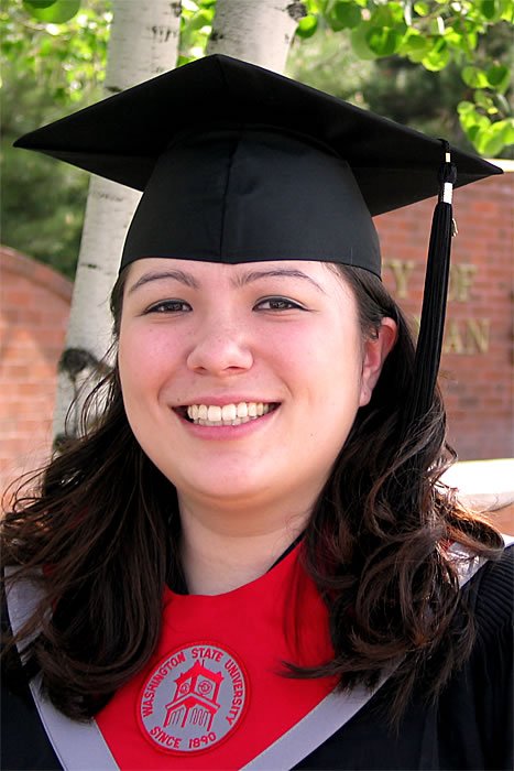 A smiling graduate wearing a cap and gown with a red shirt underneath, standing beside birch trees.