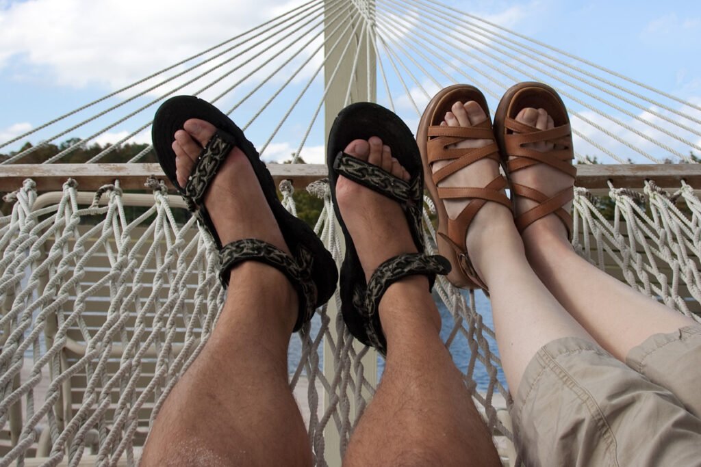 A couple's feet rest in sandals on a white mesh hammock with a bridge and blue sky in the background.