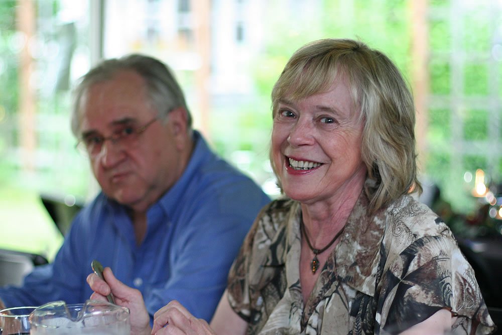 John and Kristie are seated at a table, sharing a meal. John, wearing a blue shirt, is focused on his food, while Kristie, in a patterned blouse, is smiling warmly.