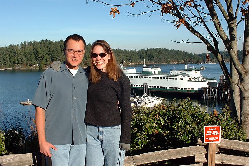 John and Kristie stand together near a scenic harbor, with boats and a forested landscape in the background. The couple smiles for the camera under clear skies.