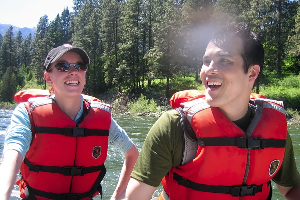 Two people wearing red life jackets smiling on a river with forest in the background.