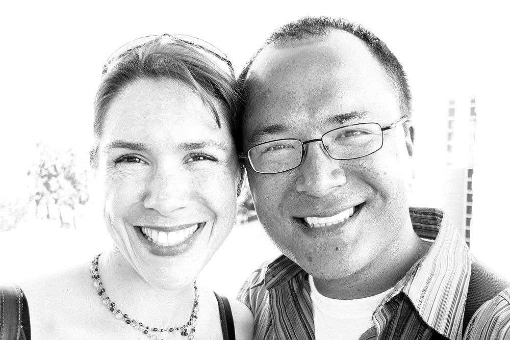 A black and white close-up photo of John and Kristie smiling, showing their faces side by side with a bright background.